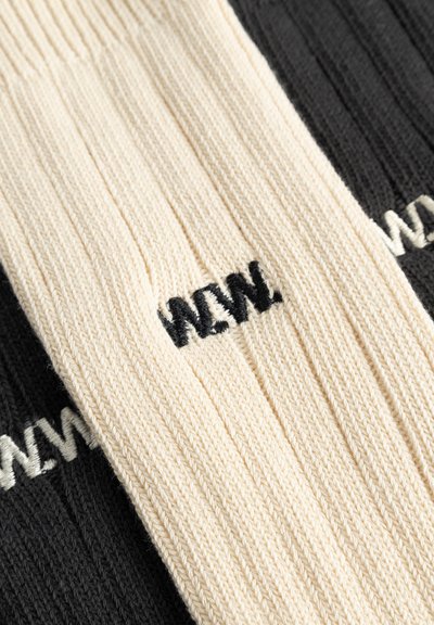 Divas pāris ribotiem zeķēm, viens krēmkrāsas ar melnu izšuvumu "MAN" un viens melns ar baltu izšuvumu "MAN", pārklājas.