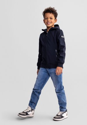 Jeune garçon souriant, marchant, portant un sweat à capuche noir zippé, un jean bleu et des baskets noires et blanches sur un fond clair uni.