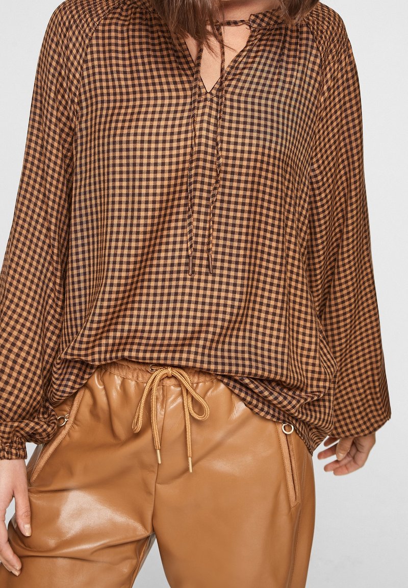 Femme portant un chemisier à manches longues à carreaux marron et noir et un pantalon marron en simili cuir avec taille à cordon.