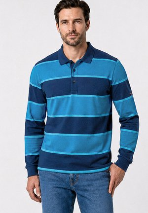 Homme portant un polo à manches longues avec de larges rayures horizontales bleues et un jean bleu, debout devant un fond uni.