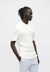 Lyhythihainen, ribbineulottu offwhite-pikeepaita klassisella kauluksella ja kaksinappisella nappilistalla, yhdistetty vaaleisiin housuihin.