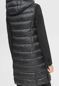 Mustavilla ilmanhihoilla varustettu untuvatakki, jossa on huppu, vuorattu pinta, vaakasaumitus ja sivutaskut. Ulkopinnassa napit.