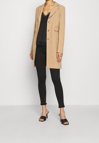 Cappotto lungo beige con tre bottoni dorati, indossato su un top a righe nere con bordo in pizzo, abbinato a jeans skinny neri e tacchi aperti.