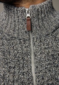 Grå strikket sweater med høj krave, der har en metallynlås og en lædertrækklap. Tekstureret mønster gennem hele stoffet.