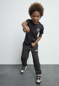 Sort T-shirt med rundt logo, sorte sweatpants med hvidt trykt logo og grå sneakers med sorte remme og accenter. Barnet laver en knytnæve.