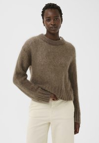 Pull tricoté marron à coupe ample, col rond et poignets côtelés. La texture semble douce et duveteuse, associé à un pantalon de couleur claire.