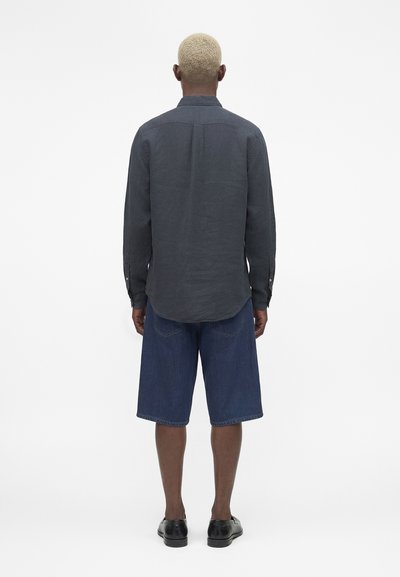 Άνδρας με κοντά ξανθά μαλλιά στέκεται πλάτης, φοράει σκούρο μακρυμάνικο μπλουζάκι, μπλε τζιν σορτς μέχρι το γόνατο και μαύρα παπούτσια.