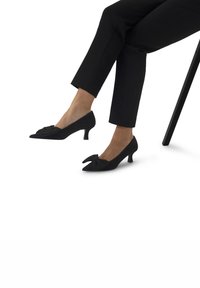 Personne assise portant un pantalon noir court et des chaussures à talons bas noires avec de grands nœuds sur fond blanc.