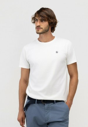 Hombre con pelo castaño ondulado, lleva una camiseta blanca lisa de manga corta y pantalones azules con un cinturón azul marino, de pie con una mano en el bolsillo.