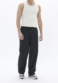 Mężczyzna w białym, bezrękawnikowym topie z prążków, w luźnych czarnych spodniach na sznurku i szaro-białych sneakersach stojący na białym tle.