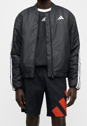 Homme portant une veste Adidas noire avec des rayures blanches et un short noir avec un motif géométrique rouge sur la jambe droite.