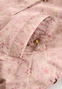 Tela rosa claro con un pequeño patrón floral y un primer plano de un bolsillo con solapa asegurado por un botón de presión de latón.