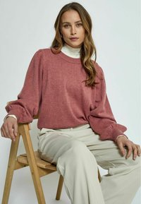 Rose-colored sweater with puffed long sleeves and ribbed cuffs, paired with white high-waisted pants, seated on a wooden chair.