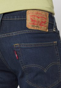 Jean bleu foncé avec des coutures jaunes, comportant une pièce en cuir à la taille arrière avec des motifs rouges et jaunes ainsi qu'une petite étiquette rouge sur la poche.