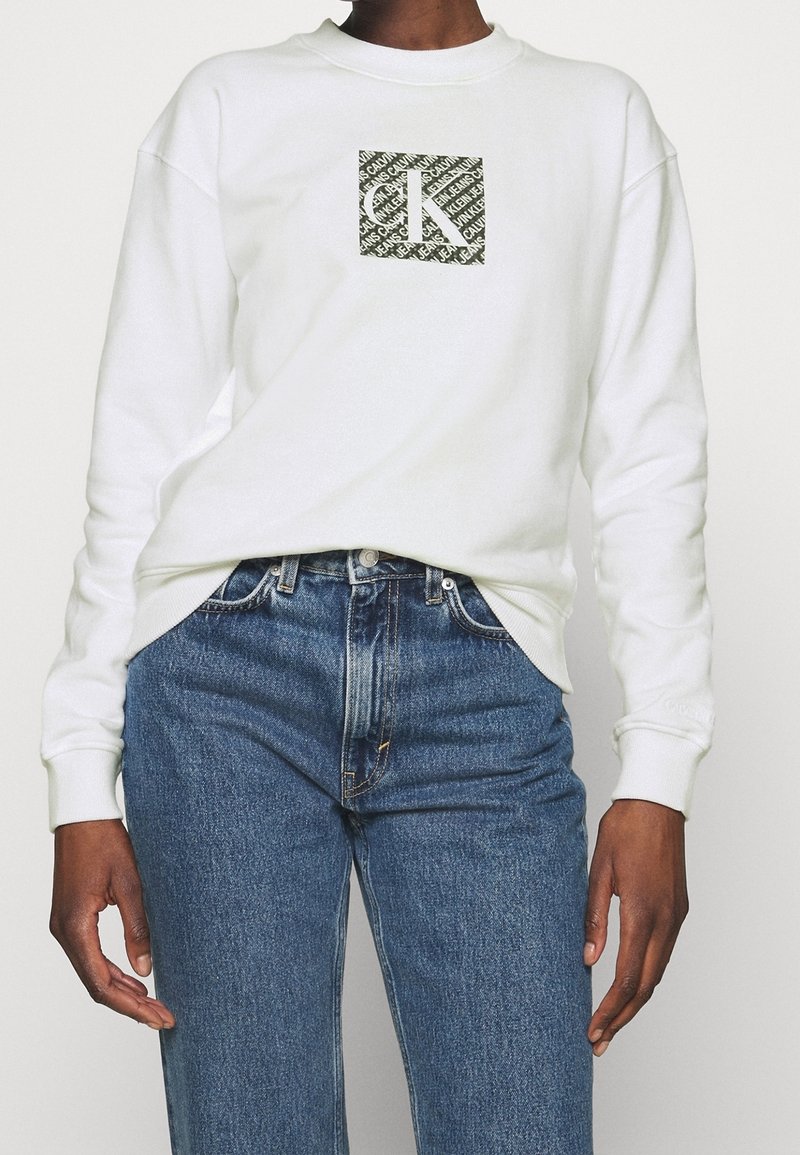 Valkoinen collegepaita, jossa on neliön muotoinen logo, jossa on vihreää kuvioitua tekstiä. Yhdistettyinä sinisiin farkkuihin, korostaen rentoa pukeutumistyylin.