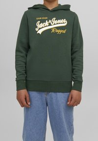 Dítě oblečené v zelené mikině s kapucí Jack & Jones a světle modrých džínách, stojící na jednobarevném šedém pozadí.