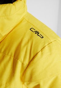 Gelbe gepolsterte Jacke mit strukturierter Oberfläche, ausgestattet mit einem Reißverschlusskragen und einem schwarzen Logo auf der Schulter. Sichtbare Nähte.