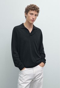 Černý dlouhý rukáv polo svetr s výstřihem do V, volný střih a manžetami, kombinovaný s bílými kalhotami na světlém pozadí.