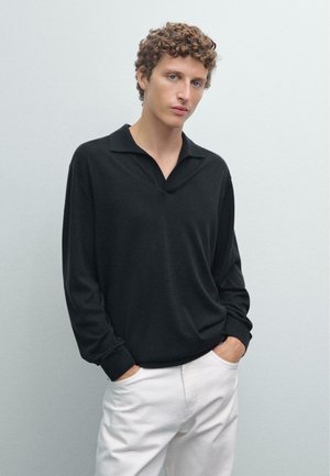 Maglione polo nero a maniche lunghe con collo a V, vestibilità ampia e maniche con polsini, abbinato a pantaloni bianchi su uno sfondo chiaro.