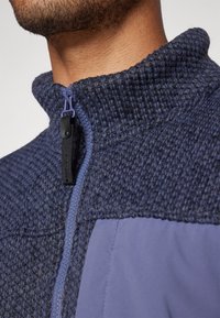 Veste zippée navy avec une section supérieure en tricot texturé et un panneau lisse en dessous. Comprend une fermeture éclair de couleur contrastante et des accents en tissu.