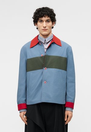 Νεαρός άνδρας με σγουρά μαλλιά που φοράει μπλε μπουφάν με κόκκινο γιακά και μανσέτες και μια σκοτεινή πράσινη οριζόντια ρίγα στο ύψος του στήθους.