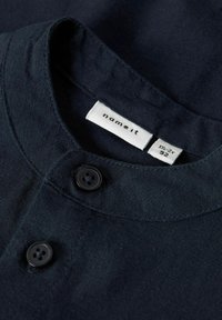 Tamsiai mėlynas medvilninis marškinėlių apsiaustas su dviem juodais mygtukais, viduje yra etiketė su užrašu „name it“ ir dydžio informacija.