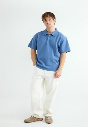 Jonge man met krullend haar, gekleed in een blauw poloshirt met korte mouwen, witte broek en beige sneakers, staand met één hand in zijn zak.