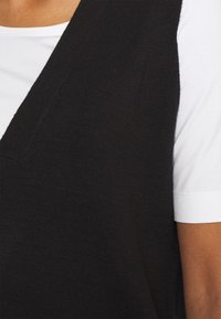 Haut sans manches en tricot noir avec un col en V, superposé sur un T-shirt blanc à manches courtes. Texture lisse et détails de style simples.