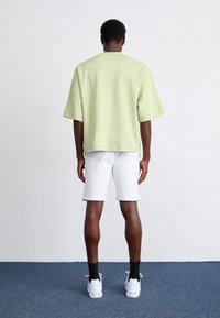 Lys grøn oversized t-shirt med korte ærmer, parret med hvide shorts. Model iført hvide sneakers og sorte sokker, vist fra ryggen.