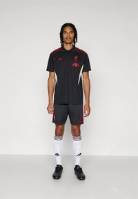 Μαύρη φανέλα της ποδοσφαιρικής ομάδας Λίβερπουλ με κόκκινες λεπτομέρειες, με γιακά και λογότυπο. Συνδυάζεται με αντίστοιχα σορτς και λευκές κάλτσες με μαύρες ρίγες.