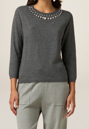 Femme portant un pull en maille gris foncé orné de perles et de pierres précieuses le long de l'encolure et un pantalon gris clair avec des poches avant.
