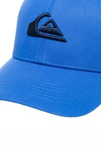 Blå keps gjord av texturerad tyg med en mörkblå broderad logotyp. Skärmen är lätt böjd med synliga sömdetaljer.