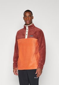 Fleecepullover med halvvinddragning, som har en rostfärgad kropp, mörkare ärmar och en beige plack med svarta knappar.