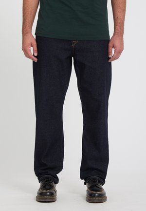 Homme portant un jean bleu foncé, des chaussures noires et une chemise vert foncé, debout devant un fond blanc uni.