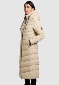 Bézs színű hosszú bélelt kabát magas gallérral, cipzáras rögzítéssel és oldalzsebekkel. Vízszintes steppelt mintázat és fekete húzózsinór díszítés.