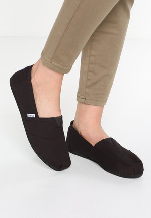 Sorte kanvas slip-on sko med en blød sål, der har en rund tå og syninger. Båret med brune halvlange bukser.