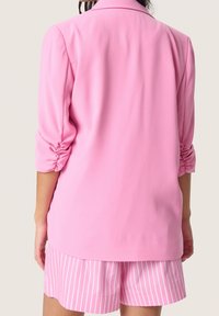 Blazer rosa con mangas 3/4 fruncidas, hecho de una tela suave. Combinado con pantalones cortos a rayas en un rosa más claro, con rayas blancas verticales.