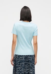 Γυναίκα  με την πλάτη γυρισμένη φοράει γαλάζιο κοντομάνικο μπλουζάκι και σκούρα μπλε φούστα με λευκά αφηρημένα γεωμετρικά σχέδια.