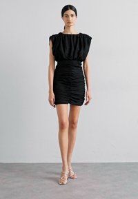 Svart mini-kjole med løs, samlet overdel og rynket skjørt. Har korte ermer og tettsittende midje. Brukt med strappy sølv sandaler.