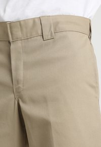 Béžové kalhoty v pase s poutky na opasek, které jsou nošeny s bílou košilí, s detailním záběrem na texturu látky a šití.