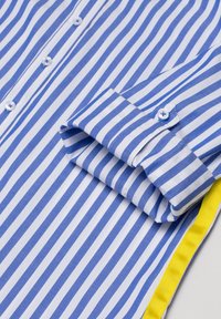 Camicia a righe blu e bianche con design a bottone, realizzata in cotone, e un accento giallo lungo l'orlo. Dettaglio con maniche arrotolate visibile.