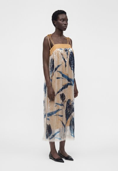 Femme portant une robe midi beige sans manches avec des motifs abstraits bleus et noirs et des ballerines noires pointues, debout devant un fond uni.