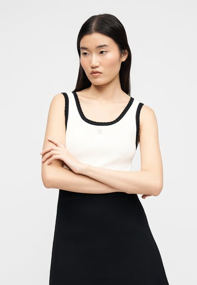 Νεαρή γυναίκα με μακριά σκούρα μαλλιά που φοράει λευκό αμάνικο τοπ με μαύρες λεπτομέρειες και μαύρη φούστα, με σταυρωμένα τα χέρια, κοιτώντας στο πλάι.