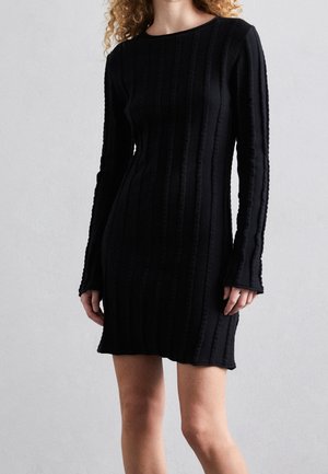Femme portant une robe noire moulante en maille côtelée à manches longues et à encolure ronde, debout devant un fond uni gris clair.