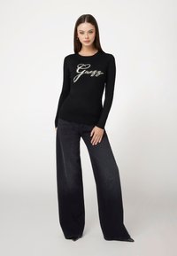 Top nero a maniche lunghe con logo metallico "Guess", abbinato a pantaloni in denim scuri a gamba larga. Materiale morbido e design aderente.