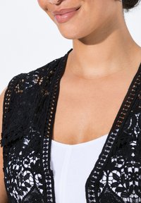 Svart spetsväst med blommönster, intrikata detaljer och rund halsringning, lager på lager över en vit topp.