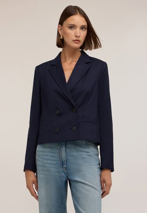 Kvinde iført en marineblå dobbeltradet blazer med guldsmykker og lyseblå jeans foran en ensfarvet beige baggrund.