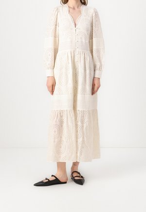 Robe longue blanche avec des motifs en œillets, à manches bouffantes, un bouton devant et une jupe à volants. Portée avec des ballerines noires à bout pointu.