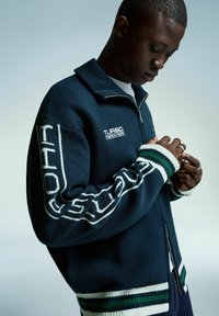 Jeune homme ajustant des bagues, portant un sweat à zip bleu marine avec des rayures blanches et vertes et le logo "Turbo Jeans" sur la poitrine et la manche.