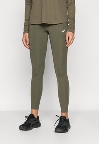 Φυστικί αθλητικό κολάν με λείο υφή και εφαρμοστή σχεδίαση· διαθέτει ένα μικρό λευκό λογότυπο στον γαστροκνήμιο. Συνδυασμένο με μαύρα αθλητικά παπούτσια.